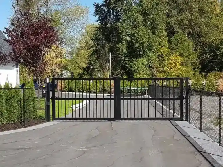 A sleek black metal sliding gate blocks a paved driveway, with ornamental gates flanked by trees and