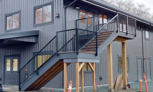 Exterior metal staircase with wooden support beams attached to a gray building. Orange traffic cones