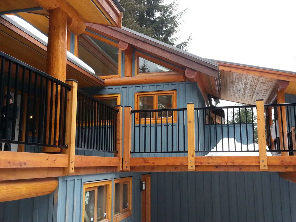 The wooden cabin exterior, enhanced by large log beams and black railings, exudes rustic charm. Its blue-gray siding contrasts beautifully with the snow-capped roof. Nestled among the trees, this serene retreat could be a perfect feature in any product gallery.