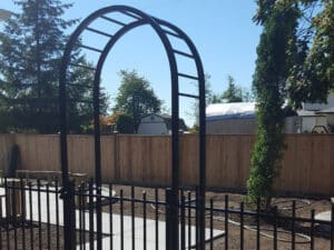 Black metal garden arbor and fence in a yard with bare soil, concrete walkway, wooden privacy fence, and trees in the background—perfect for pairing with durable Aluminum Railings Langley for added style and security.