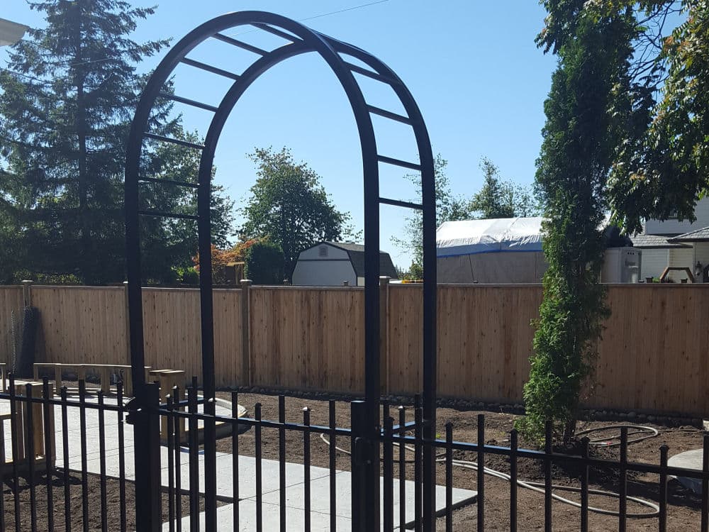 Black metal garden arbor and fence in a yard with bare soil, concrete walkway, wooden privacy fence, and trees in the background—perfect for pairing with durable Aluminum Railings Langley for added style and security.