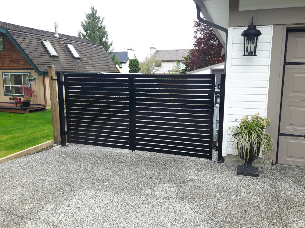 Black metal gate with horizontal slats on a gravel driveway, next to a white house and potted plant, with residential houses and greenery in the background.