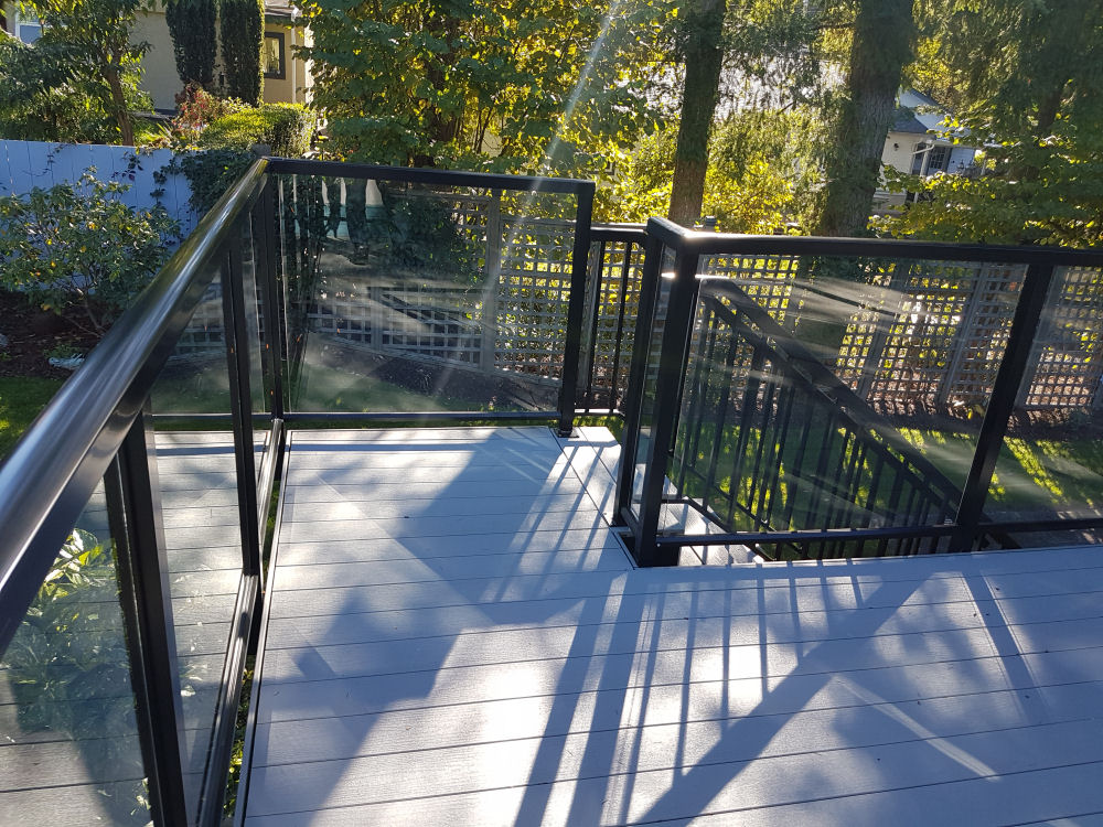 Modern outdoor deck with glass and metal railing, overlooking a garden with trees and bushes. Sunlight creates shadows on the deck floor.
