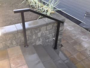 A black metal handrail is installed along concrete steps next to a cinder block retaining wall, with paving stones and tall plants nearby—creating a sleek alternative to traditional aluminum railings.