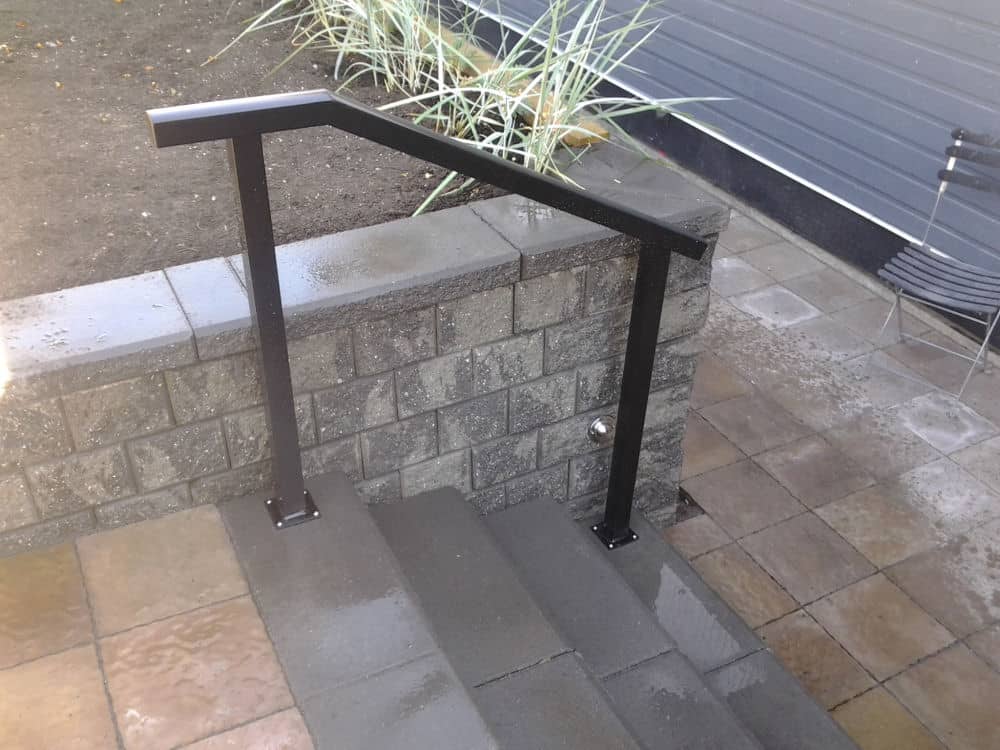 A black metal handrail is installed along concrete steps next to a cinder block retaining wall, with paving stones and tall plants nearby—creating a sleek alternative to traditional aluminum railings.