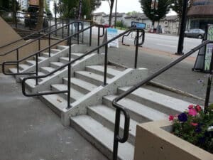 Concrete stairs with multiple handrails alongside a sidewalk. Planters with flowers are visible on the right.