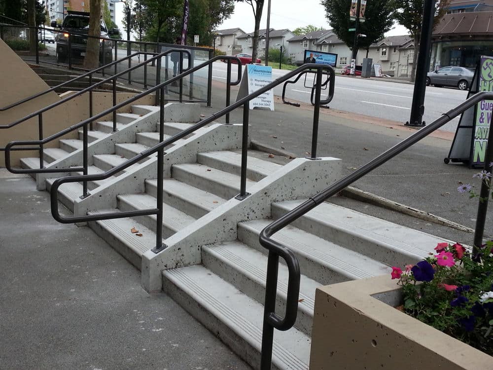Concrete stairs with multiple handrails alongside a sidewalk. Planters with flowers are visible on the right.