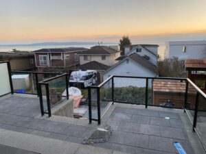 Modern patio with glass and metal railing by a skilled Metal Railing Contractor overlooks residential rooftops and the water at sunset, with construction materials and equipment visible on the lower level.