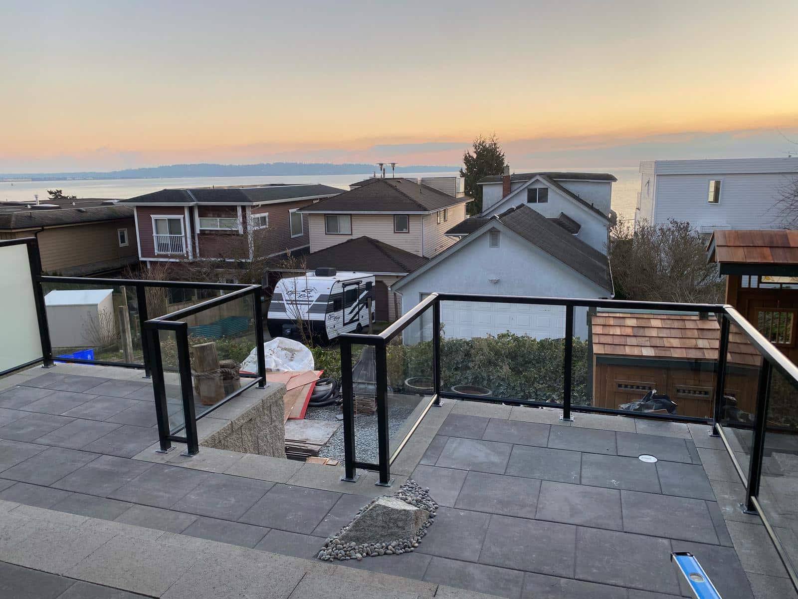Rooftop patio with sleek railings overlooking a charming residential area and a distant sea view during sunset. Paved flooring and a small rock garden complete the serene ambiance.