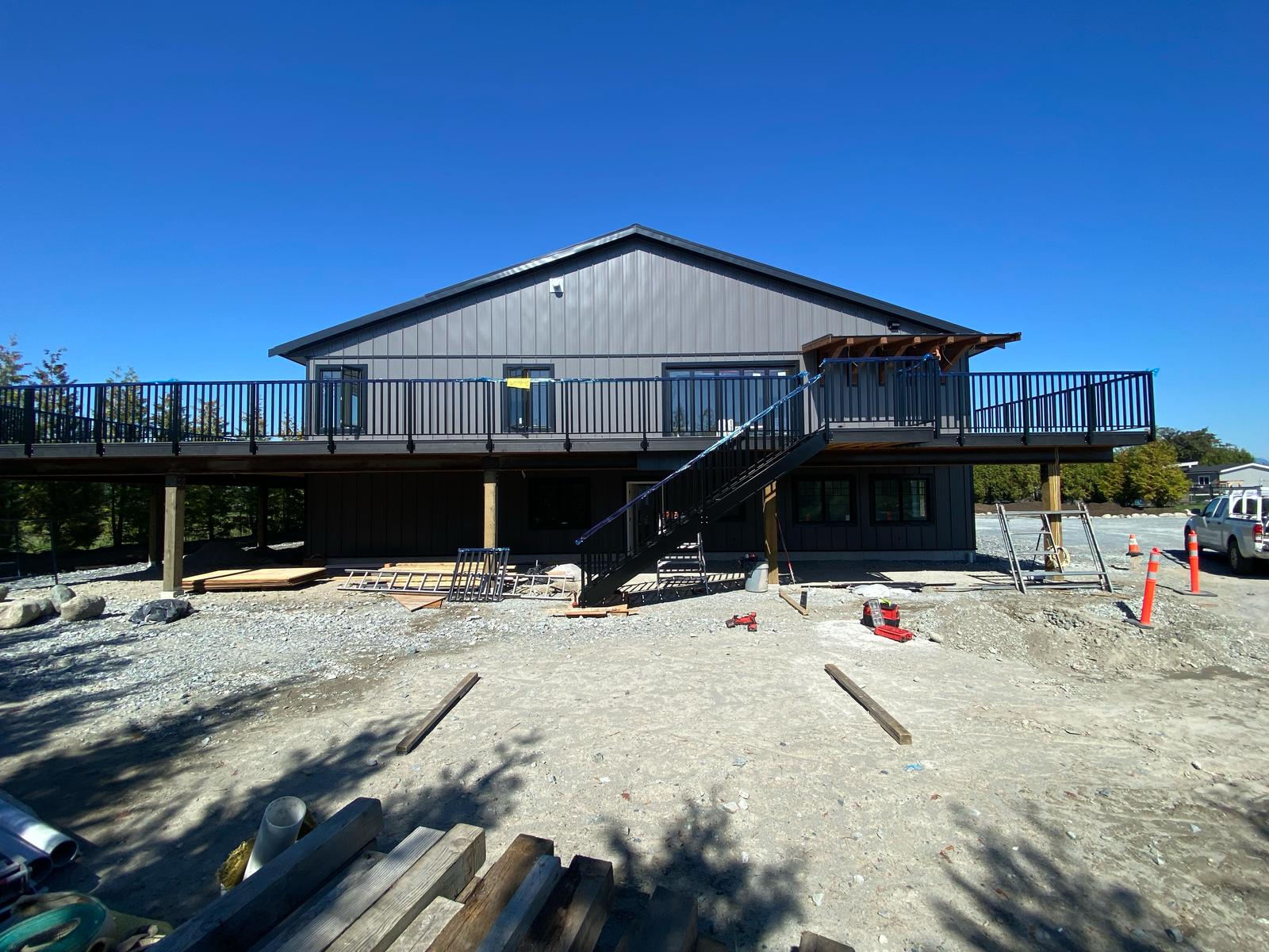 A two-story building with a large wraparound deck featuring picket railings, an exterior staircase, and gravel lot under a clear blue sky; construction materials are scattered in the foreground.
