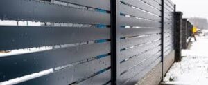 Close-up view of a modern horizontal metal slat fence outdoors, featuring sleek aluminum railings with water droplets on them and snow covering the ground.