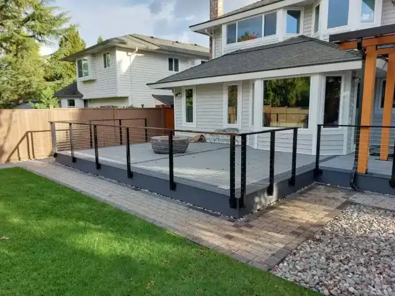 Backyard view of a modern house with a raised deck enclosed by sleek black aluminum railings, bordered by grass, pavers, and decorative rocks.