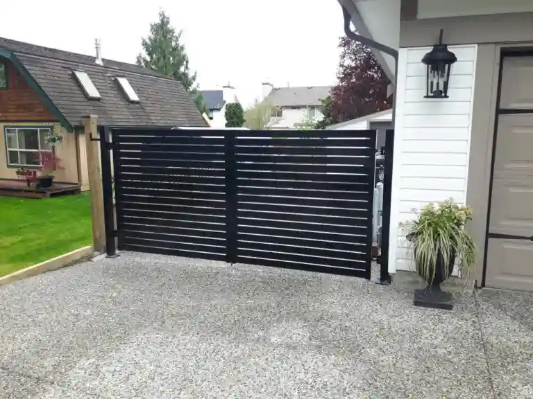 A modern black metal horizontal slat gate, complemented by elegant Aluminum Railings Langley, blocks the driveway beside a white house with a potted plant and outdoor lantern.