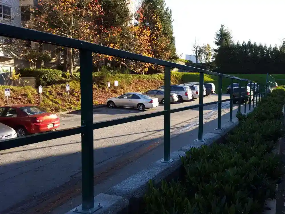 A row of parked cars lines a street next to a building, with outdoor railings and bushes in the foreground under a clear sky.