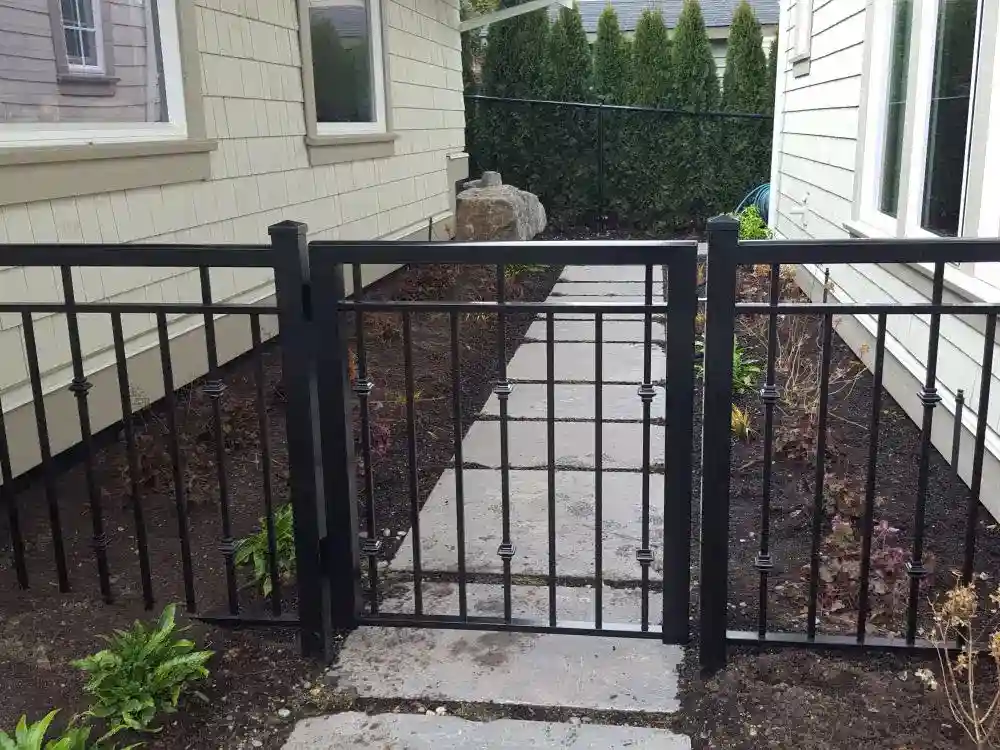 A black metal gate blocks a stone pathway between two beige houses, with sparse plants and soil on either side of the path. If you’re considering alternatives like aluminum railing, exploring Aluminum Railing Pricing can help you find the perfect balance of style and budget for your outdoor space.