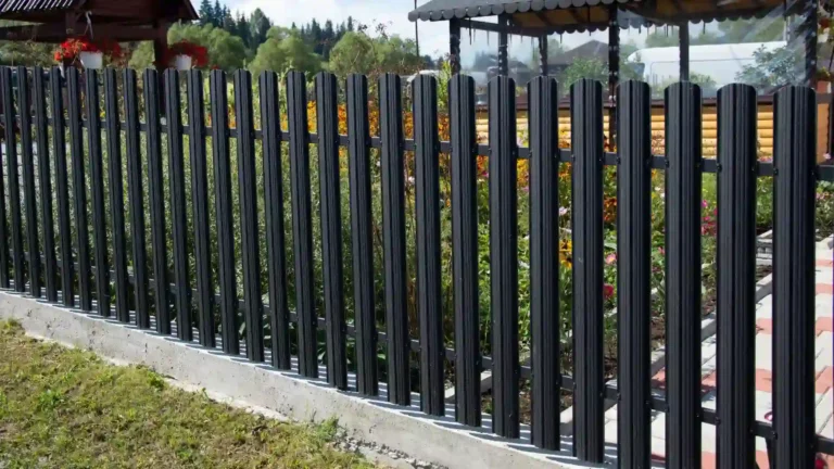 A tall black metal fence with vertical slats stands on a concrete base, featuring sturdy metal railings that enclose a garden with green plants and flowers. A wooden structure and trees are visible in the background.