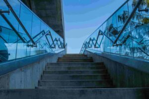 Concrete staircase with glass and metal railings, leading upward toward a blue sky, with trees partially visible on the right side—showcasing What Cable Glass Railings Options in Langley Township can offer for modern outdoor spaces.