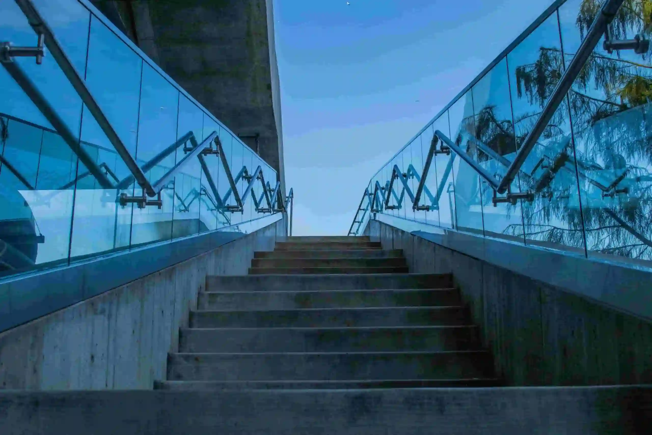 Concrete stairs lead upward, bordered by sleek glass railings with metal supports, under an open sky with a building structure partially visible to the left.