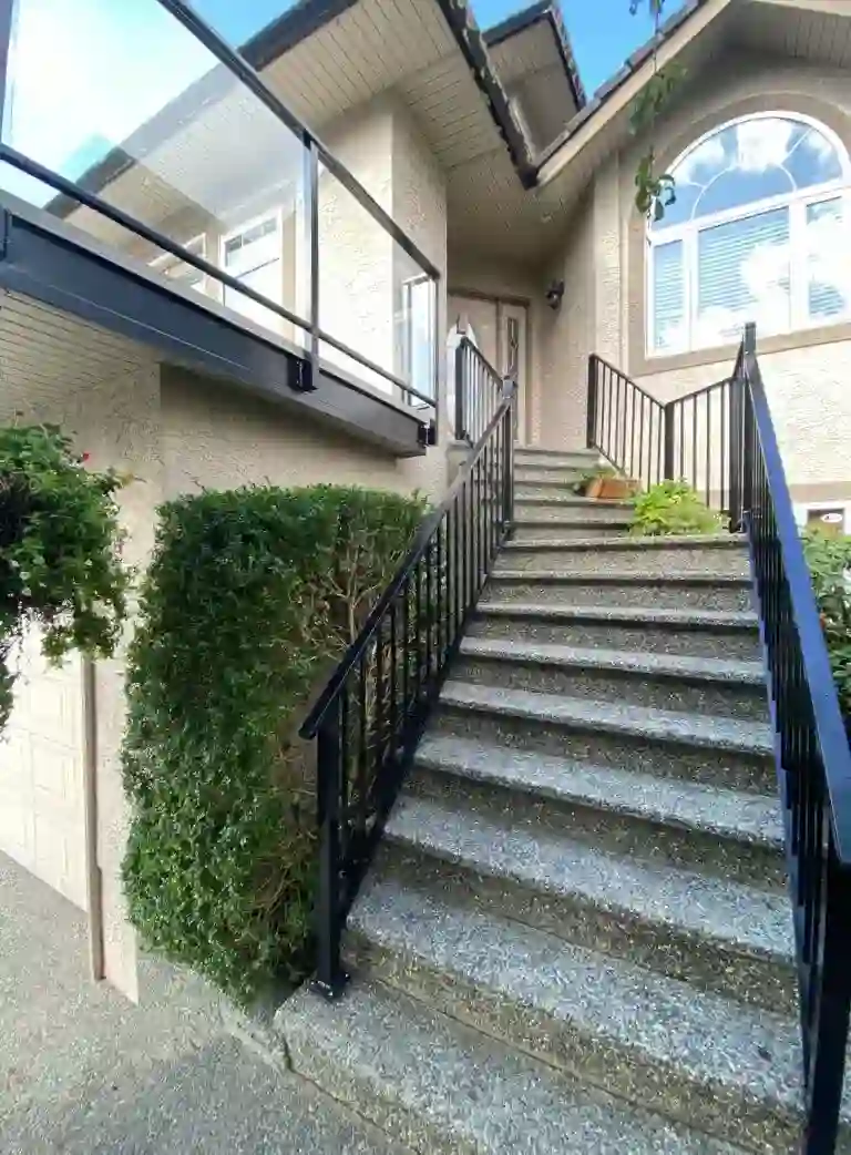 Concrete steps with a sleek metal railing lead up to the front door of a beige house, featuring a large arched window and glass balcony above.