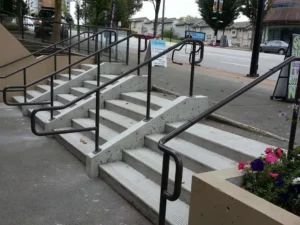 Concrete stairs with five steps and four black metal handrails, leading from a sidewalk up to a higher level beside a street, with flowers and buildings visible in the background. The sturdy railings reflect careful planning, meeting the requirements for metal railing installation Langley residents follow, including securing the proper permit for metal railing installation.