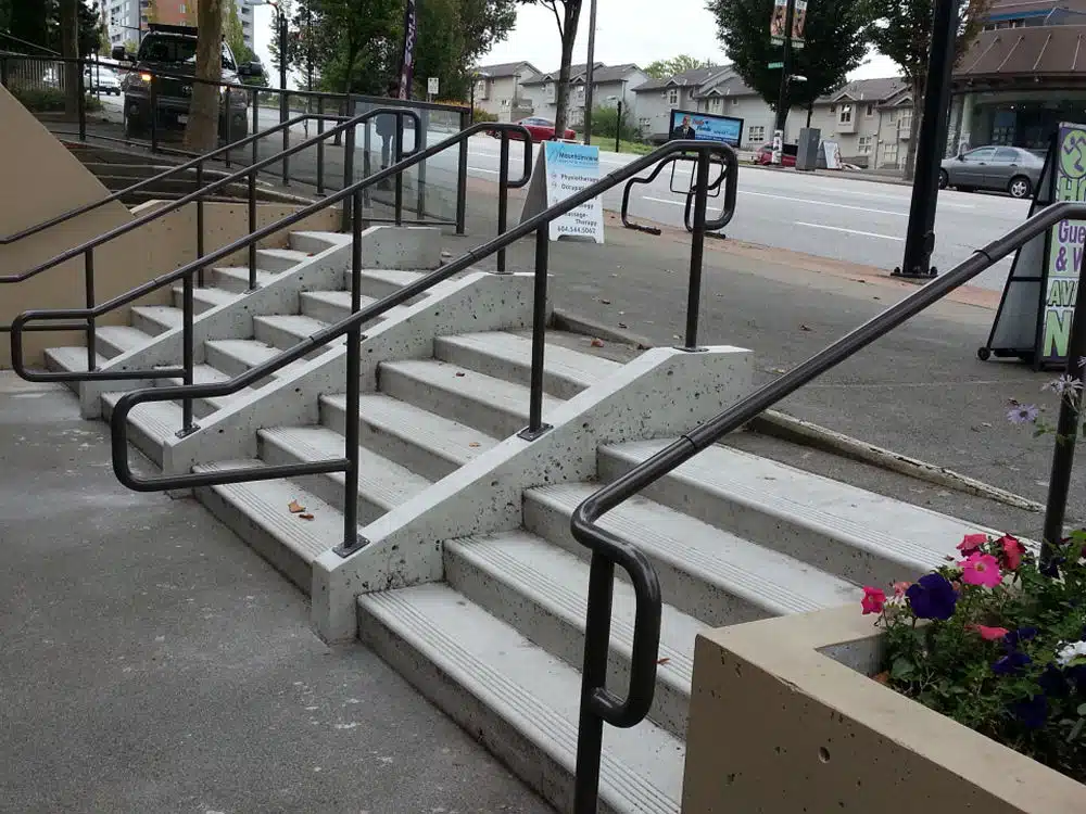Concrete stairs with five steps and four black metal handrails, leading from a sidewalk up to a higher level beside a street, with flowers and buildings visible in the background. The sturdy railings reflect careful planning, meeting the requirements for metal railing installation Langley residents follow, including securing the proper permit for metal railing installation.