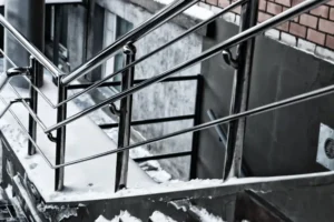 Snow-covered outdoor concrete steps with sturdy aluminum railings installed on stairs, next to a brick building. Snow is piled on the edges, and the ground below is also partially covered in snow.