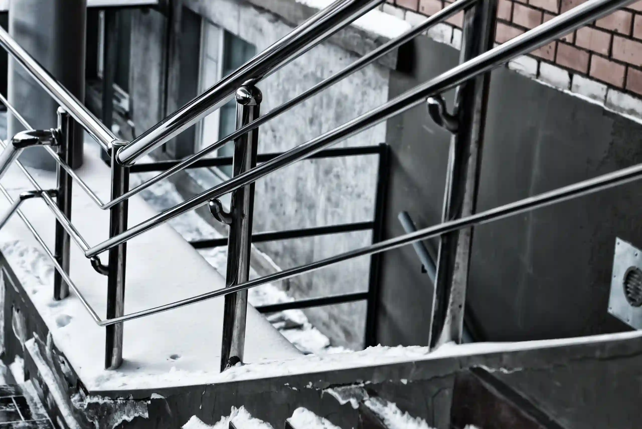 Snow-covered outdoor concrete steps with sturdy aluminum railings installed on stairs, next to a brick building. Snow is piled on the edges, and the ground below is also partially covered in snow.