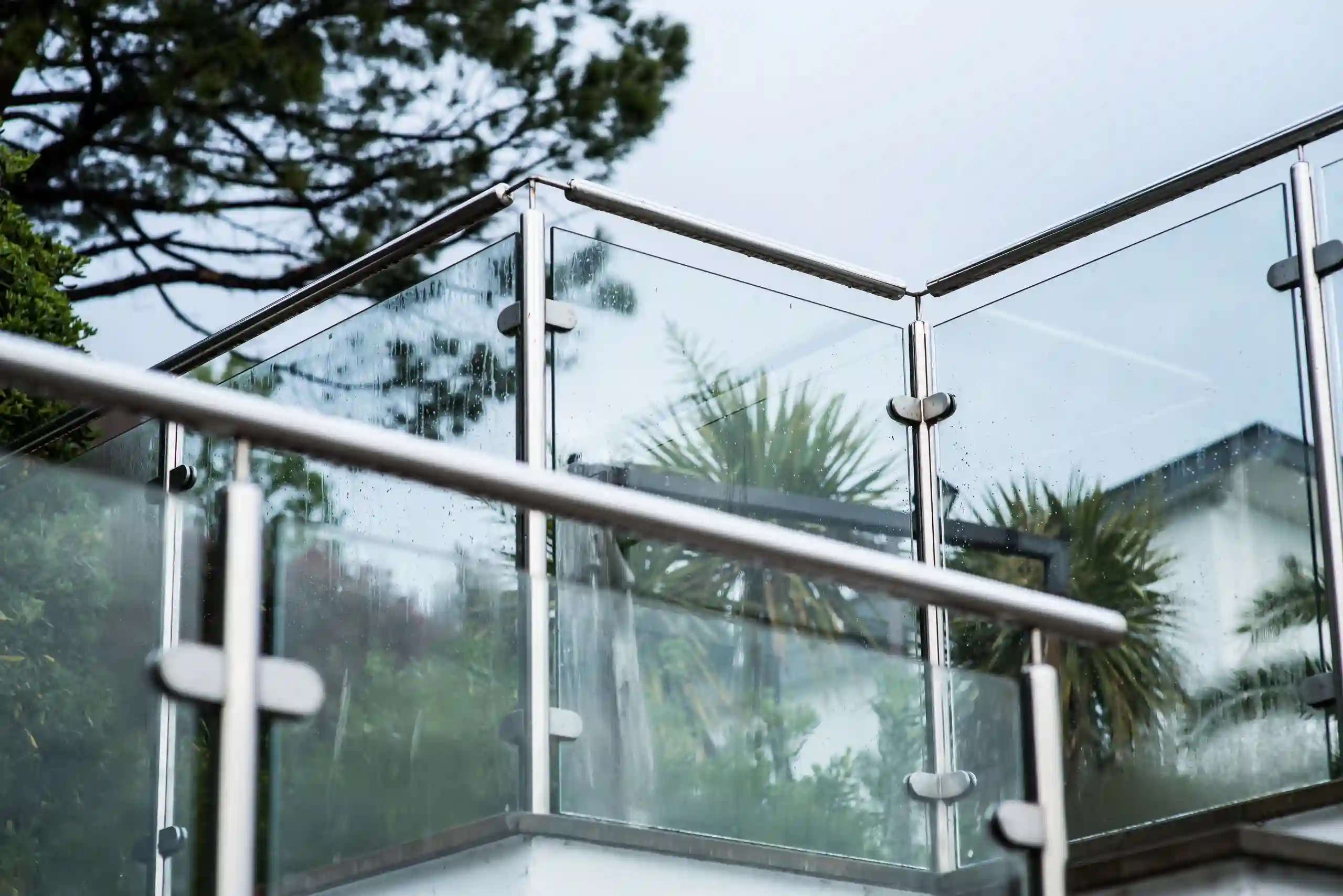 Modern glass and metal balcony railing with raindrops, set against greenery and a cloudy sky.