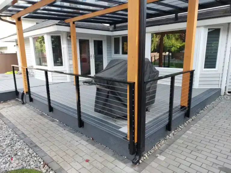 Modern backyard deck with a glass roof, custom aluminum railings, and a covered grill at the center, attached to a gray and white house with a surrounding paved walkway and rocks.