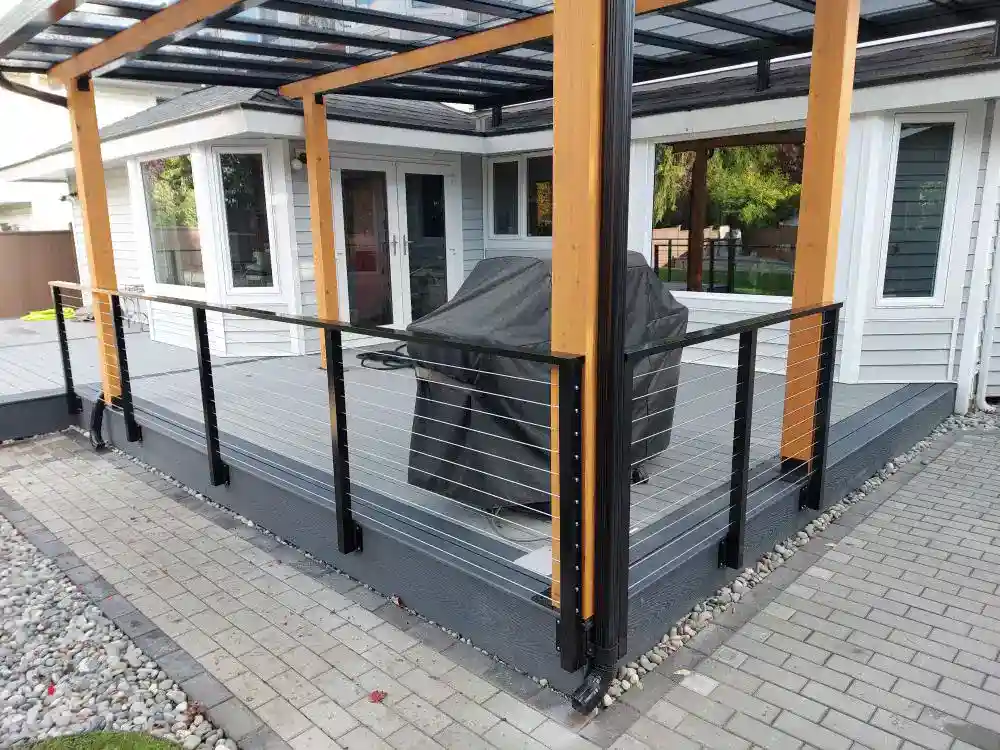 Modern backyard deck with a glass roof, custom aluminum railings, and a covered grill at the center, attached to a gray and white house with a surrounding paved walkway and rocks.