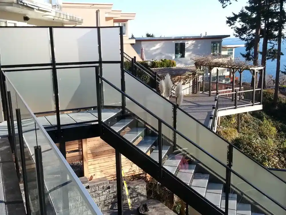 Outdoor glass-panel stairway with sleek black metal framing and premium Aluminum Railings North Vancouver, leading down to a wooden deck and gazebo overlooking trees and water in the background.