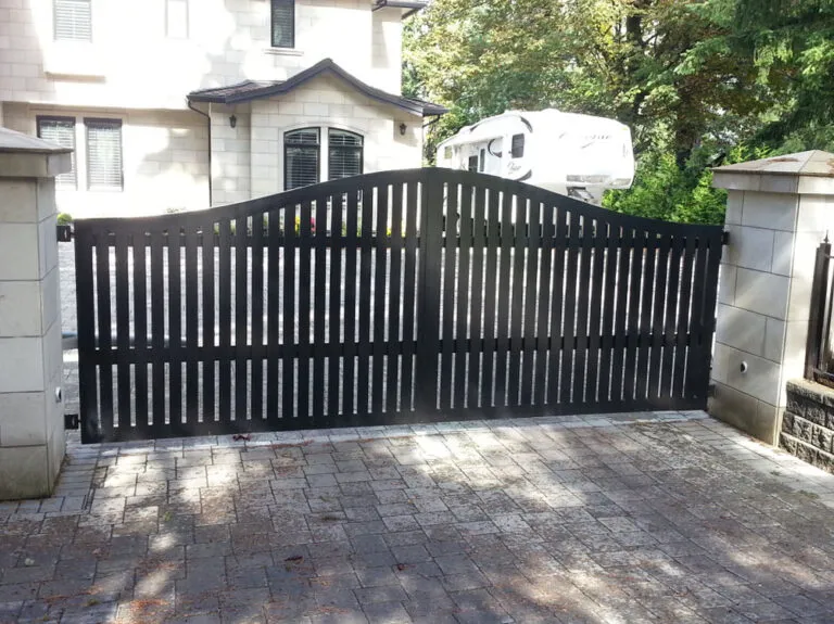 A black metal driveway gate with vertical slats is closed between two stone pillars in front of a house, with a white RV visible in the background, showcasing one of the most stylish and secure gates available.