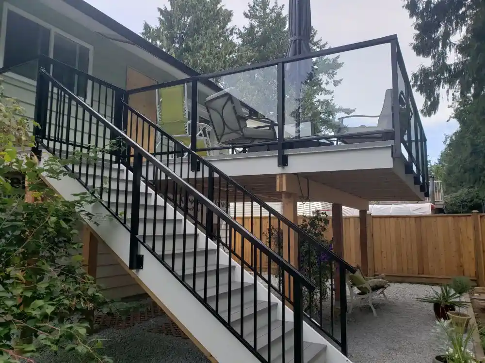 Outdoor staircase with black metal railings leads to a raised deck featuring elegant glass panels by Glass Railings Coquitlam, complemented by patio furniture and an umbrella; fenced yard with trees and gravel below.