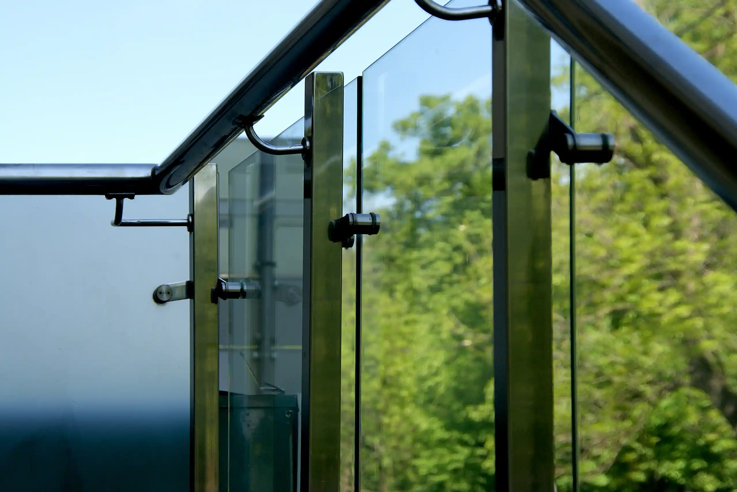 Close-up view of glass panels and metal supports on a structure with trees and blue sky visible in the background.
