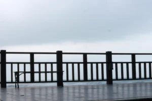 Wooden deck with aluminum railings overlooks a misty, overcast horizon. A bench sits on the left side, and the floor appears wet, suggesting recent rain. The aluminum railing safety features provide added peace of mind while enjoying the view.