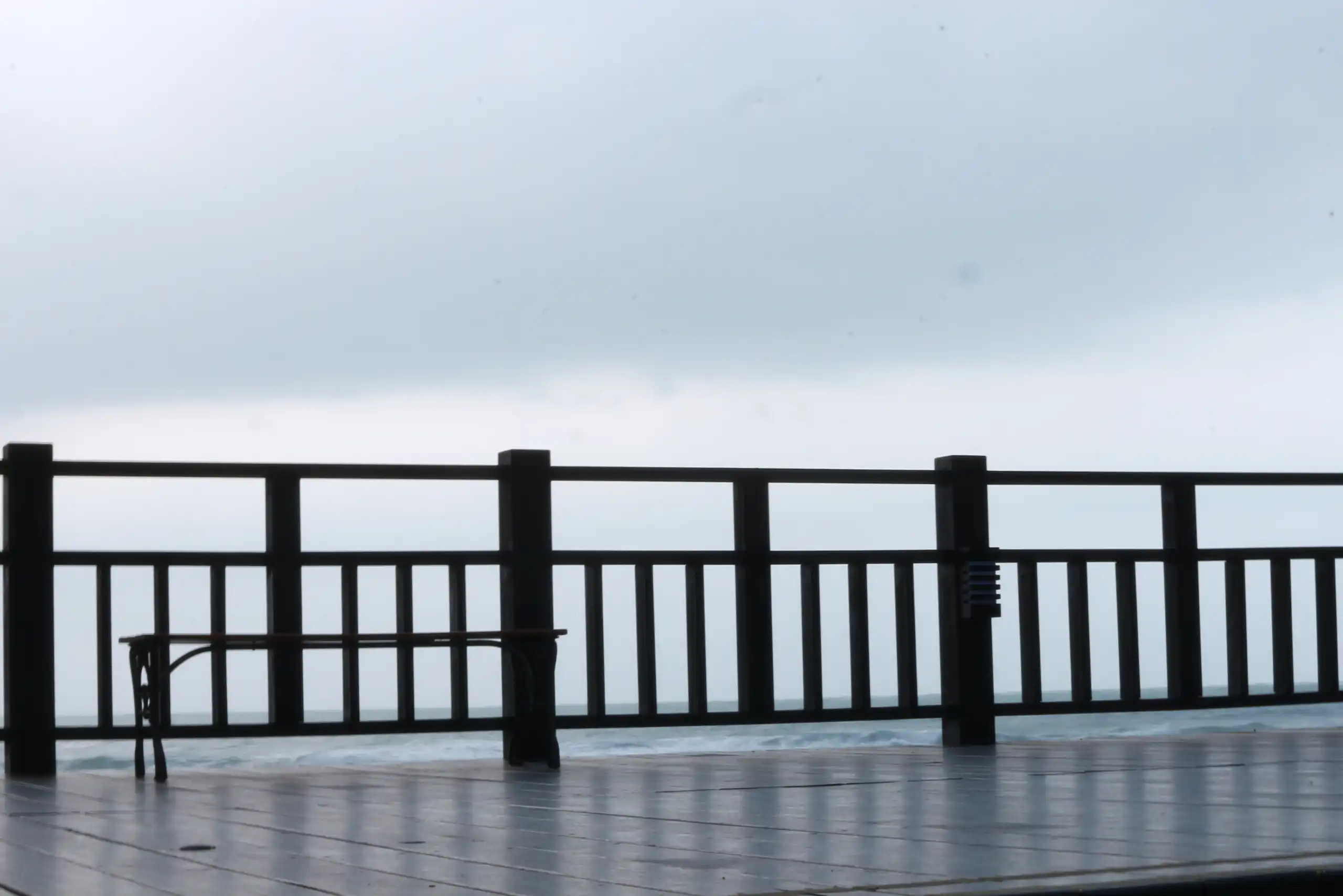 Wooden deck with aluminum railings overlooks a misty, overcast horizon. A bench sits on the left side, and the floor appears wet, suggesting recent rain. The aluminum railing safety features provide added peace of mind while enjoying the view.