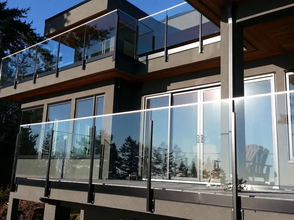 Modern two-story house featuring large glass balcony railings by LDH Aluminum Ltd. – Railings & Gates, with multiple sliding glass doors reflecting trees and sky in the background.