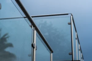 Close-up of a modern glass and metal balcony railing against a cloudy blue sky, with faint reflections of trees visible in the glass panels—an elegant example of the many types of glass railings that combine style with unobstructed views.