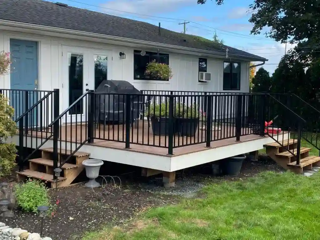 A wooden deck with black metal railings—crafted in the style of Metal Railings New Westminster—is attached to a white house, featuring stairs on both sides and a grill, chairs, and potted plants on the deck.