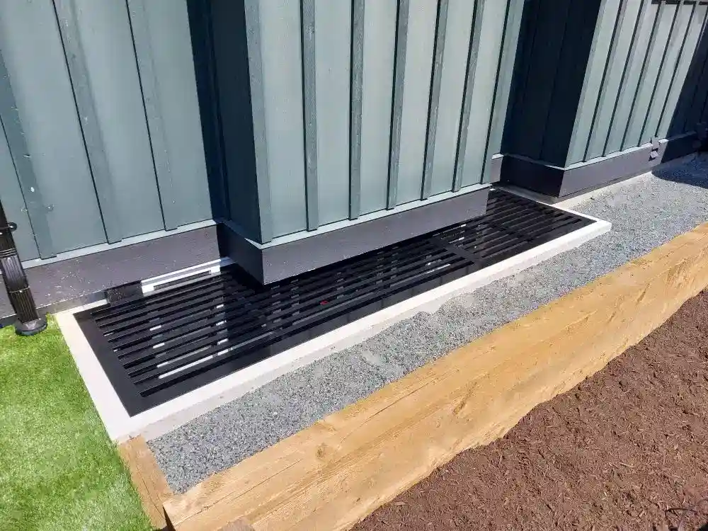 A black metal grate covers a drainage area next to a building with green siding, bordered by concrete, wood, grass, and soil—demonstrating how well-designed drainage solutions and related services contribute to the upkeep of exterior spaces.