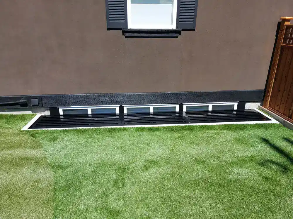 Four small horizontal windows at the base of a brown building, protected by sturdy black window grates, bordered by artificial grass.