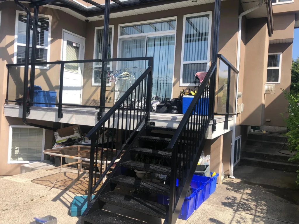 A back porch with black metal railings resembling the sleek style of aluminum fencing Abbotsford is known for, various household items, and blue storage bins sits outside a beige house on a sunny day.
