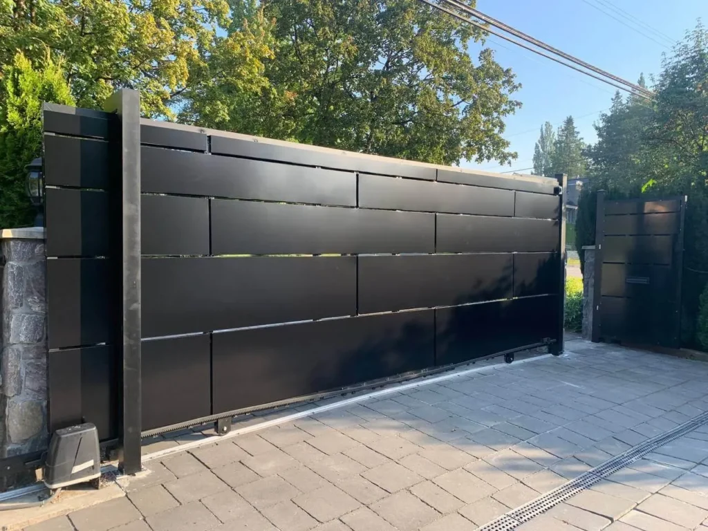A large black metal sliding gate is installed on a stone driveway, complemented by sleek aluminum fencing Abbotsford residents trust for both security and style, with trees and greenery visible in the background.
